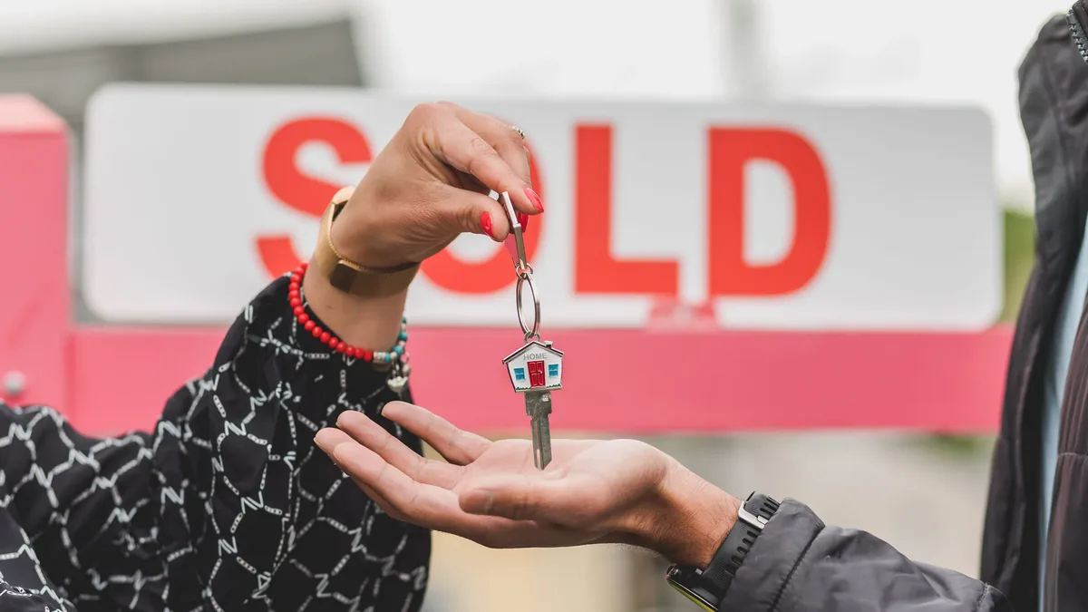 A person handing over a set of keys to another person in front of a "SOLD" sign.